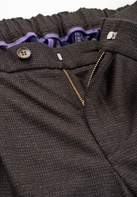 Pantalon marron foncé texturé avec une fermeture éclair dorée partiellement ouverte, un bouton supérieur et une doublure en tissu violet visible à l'intérieur de la ceinture.