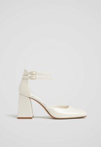 Escarpins à talons hauts en cuir verni blanc avec un bout rond, un talon carré et deux brides ajustables à la cheville. Texture lisse et design minimaliste.