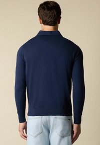 Námořnická modrá polo košile s dlouhým rukávem a límečkem. Pletená textura a žebrované manžety. V kombinaci se světle modrými džíny s zadními kapsami.