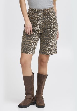 Donna che indossa pantaloncini leopardati fino al ginocchio, top grigio e stivali di pelle marroni fino al ginocchio, in piedi davanti a uno sfondo semplice.