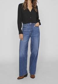 Chemise noire à manches longues avec boutons, associée à un jean bleu à jambes larges. Le jean présente une taille haute et un léger délavage. Des bottines marron complètent le look.