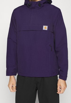 Veste imperméable violette avec capuchon, poche avant et cordon de serrage. Caractéristiques d'un patch logo et de poignets élastiques. Tissu texturé, design décontracté.