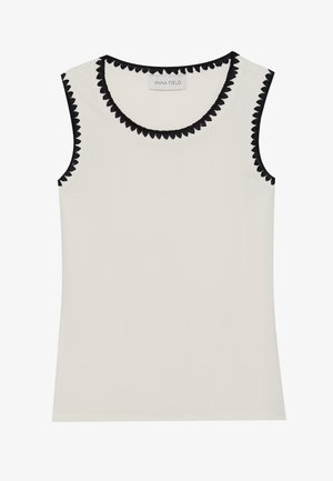 Bezrukava kremasta majica s rebrastom teksturom, ukrašena crnim valovitim obrubom oko izreza i rukava. Oznaka glasi "ANNA FIELD."