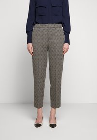 Žena oblečená v šedých kostkovaných zúžených kalhotách, námořnické haleně s dlouhým rukávem a bílých lodičkách s černými špičkami, stojící na jednobarevném pozadí.