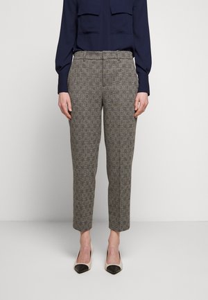 Kvinde iført gråternet taperede bukser, marineblå langærmet bluse og hvide hæle med sorte tådæksler, stående mod en ensfarvet baggrund.