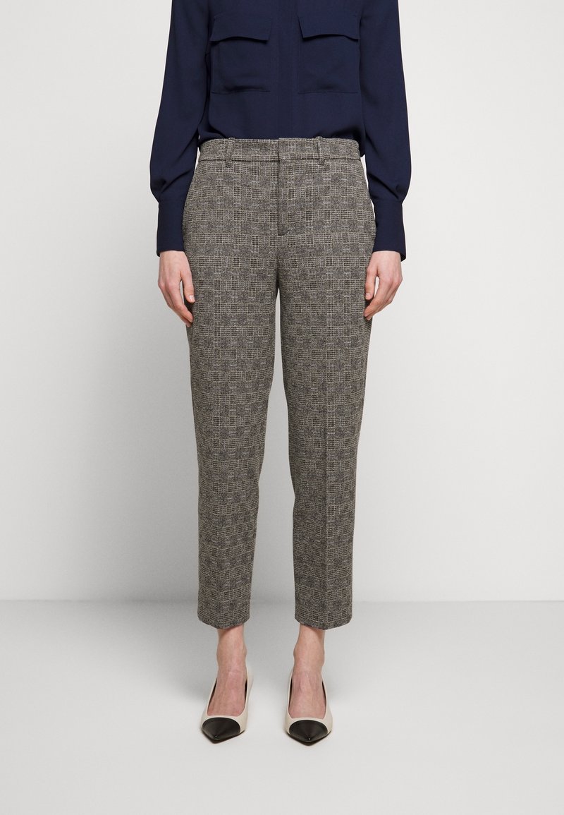 Žena oblečená v šedých kostkovaných zúžených kalhotách, námořnické haleně s dlouhým rukávem a bílých lodičkách s černými špičkami, stojící na jednobarevném pozadí.