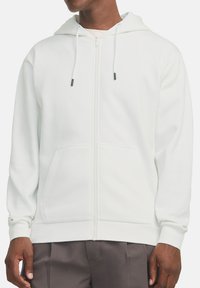 Hoodie blanc zippé en matériau doux, avec une capuche doublée, des cordons de serrage et des poches avant. Texture lisse avec des poignets côtelés.
