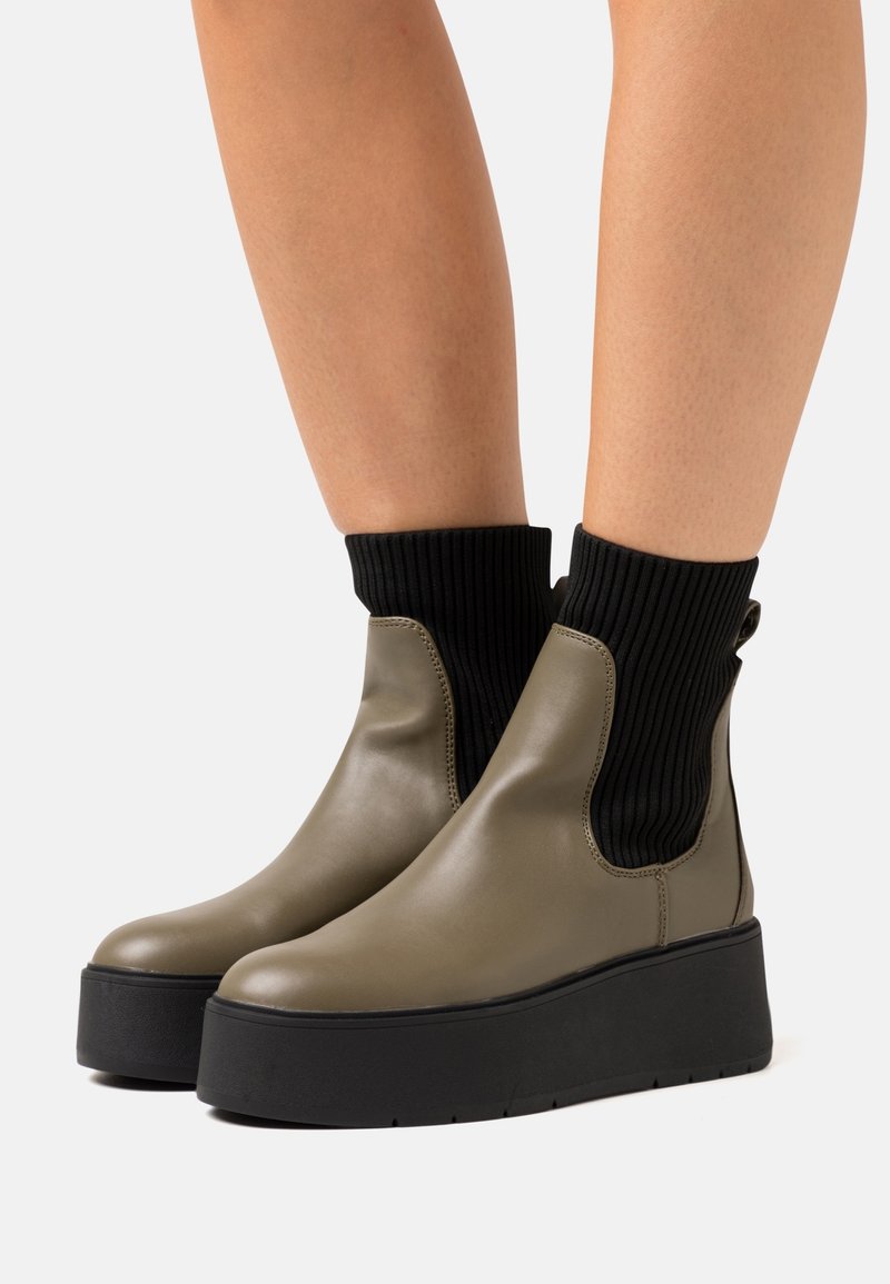 Bottines vert olive avec un extérieur en cuir lisse, un col en maille noir ondulé, et une semelle plateforme noire épaisse.