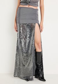 Falda gris de lentejuelas con una gran abertura en un lado, combinada con botas negras hasta la rodilla. Textura lisa y reflectante, diseño ajustado.