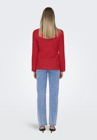 Rød tætsiddende blazer med lange ærmer, tekstureret stof, slids bagpå, kombineret med lyseblå jeans med lige ben og sorte spidse hæle.