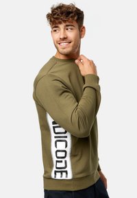Sweatshirt vert olive avec un col rond, des poignets côtelés et un panneau latéral blanc portant un texte noir audacieux. Tissu doux et design ajusté.