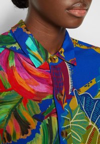 Primo piano di una persona che indossa una camicia blu vivace con motivi floreali tropicali multicolori e bottoni marroni.
