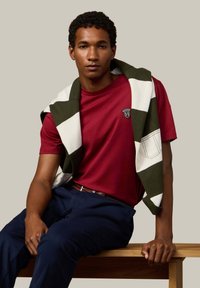 Rode katoenen t-shirt met een logo, marineblauwe broek en een gebreide groen-witgestreepte trui die over de schouders is geslagen. Houten bank op de achtergrond.