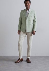 Ljusgrön dubbelknäppt blazer i linne, parat med en blommig skjorta med knappar och ljusbeiga byxor, kompletterad med bruna läderskor.