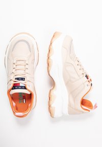 Scarpe da ginnastica beige con tomaia testurizzata, soletta arancione e suola color crema. Le caratteristiche includono lacci e un'etichetta con il logo sulla linguetta.