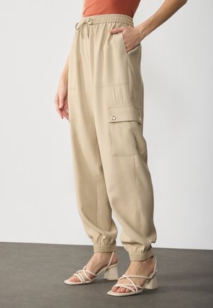 Beige cargobroek met elastische taille en boorden, zijflappenzak met knoop, gedragen met beige sandalen met blokhak en bandjes.