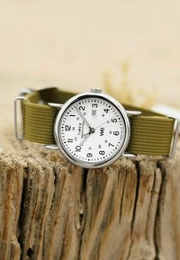 Timex Weekender kell valge puidust numbrite ja kuupäevanäidikuga, millel on mustad numbrid ja oliiviroheline kangast rihm, puhkab kulunud puidust pinnal.