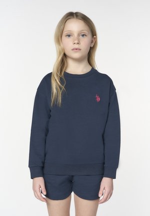 Ragazza giovane con lunghi capelli biondi che indossa una felpa blu navy con logo ricamato rosa e pantaloncini blu navy abbinati, in piedi davanti a uno sfondo bianco.