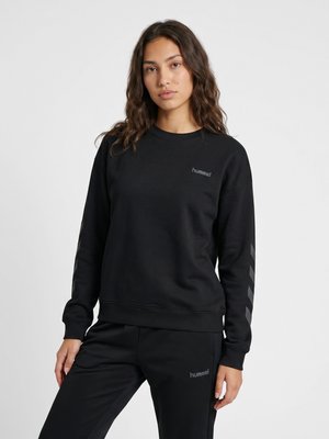 Kvinde med langt brunt hår iført en sort hummel sweatshirt og matchende sorte bukser mod en ensartet lys baggrund.