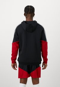 Sweatshirt com capuz, corpo preto e mangas vermelhas, apresentando riscas brancas de lado. Usado sobre calções pretos e vermelhos, com textura em malha visível.