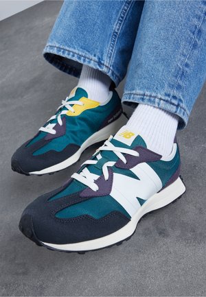 Pieds portant des baskets New Balance bleues, noires et blanches avec des chaussettes blanches et un jean bleu, debout sur un sol en béton gris.