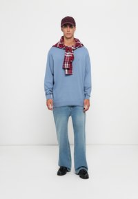 Felpa blu in cotone, camicia a quadri rossa e bianca legata attorno al collo, jeans a zampa d'elefante in lavaggio chiaro e scarpe nere, abbinati a un cappellino scuro.