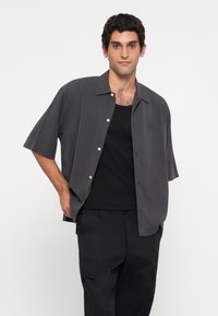 Cămașă cu mânecă scurtă, de culoare gri închis, cu un croi lejer, cu nasturi pe față și o textură fină, purtată peste un top negru și pantaloni cargo.