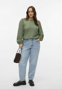 Maglietta lunga a maniche puff con texture verde, abbinata a jeans a vita alta blu chiaro e scarpe stringate nere, mentre tiene una borsa marrone.