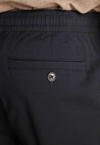 Los pantalones navy presentan una cintura elástica y un bolsillo trasero con un botón plateado. La tela parece suave con una sutil textura.