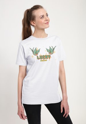 Kvinde med brunt hår iført en hvid t-shirt med to grønne fugle og teksten "Lucky sailor", står foran en ensfarvet lys baggrund.