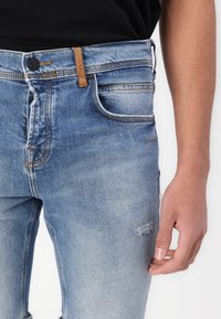 Svetlo modri jeans z ožjim krojem, z gumbom za zapiranje, kontrastnim šivanjem in manjšimi obrabljenimi detajli na kolenu.