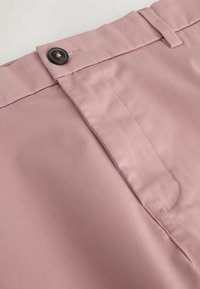 Gros plan sur la taille d'un pantalon rose blush avec un bouton noir, une passoire de ceinture et un détail de braguette cousu sur un tissu lisse.