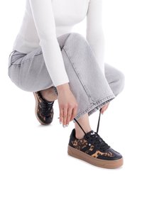 Zapatillas de leopardo en negro y marrón con suela de goma, cordones planos y textura suave, combinadas con jeans de pierna ancha en color gris.