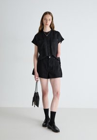 Juodas trumpomis rankovėmis marškiniai su mygtukais, derinami su juodomis šortais. Modelis laiko mažą juodą krepšį, vilkėdamas juodas kojines ir loafers batus.