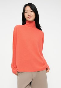 Ung kvinne med langt svart hår iført en korallfarget høyhalset genser og beige bukser, stående foran en hvit bakgrunn.