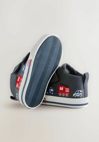 Bērnu tumši zilas augstas kurpes ar izšūtām ārkārtas transportlīdzekļu ilustrācijām un strukturālām baltām zolēm, novietotas uz bežīga fona.