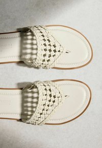 Paire de sandales blanc cassé avec des brides tressées, des boucles pour les orteils et des semelles marron clair sur une surface texturée claire.