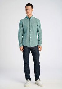 Ljusgrön skjorta med knappar i en texturerad tyg, kombinerad med mörkblå jeans och vita skor, stående mot en enkel bakgrund.