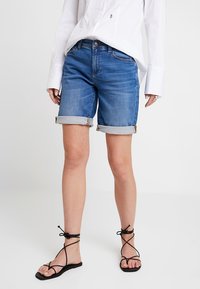 Denim shorts in blue with rolled cuffs, featuring a classic five-pocket design and a button closure. Paired with black strappy sandals.