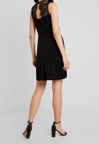 Mustat olkaimettomat mekot, joissa on pitsinen selkäosa, A-linjainen muoto, sileä pinta, polvipituus, yhdistetty mustien korkokenkien kanssa.