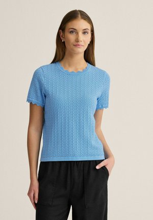 Ung kvinde iført en lyseblå strikket top med korte ærmer med bølgede kanter og sorte bukser, stående med den ene hånd i lommen.