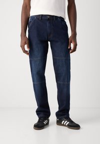Dark blue denim jeans featuring a straight-leg cut, contrast stitching, and patchwork design. Worn with black sneakers featuring white stripes.