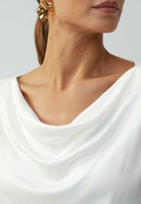 Weißes Top mit Drapé-Ausschnitt und weicher Textur. Goldfarbene, abstrakt geformte Ohrringe betonen den Look. Nahaufnahme von Hals und Schultern.