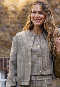 Beżowa kurtka z tkaniny tweed z złotymi guzikami, fakturowany materiał, luźny krój; zestawiona z pasującą beżową bluzką i spodniami z tkaniny tweed.