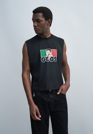 Άνδρας που φοράει μαύρο αμάνικο μπλουζάκι με χαρακτήρα κινουμένων σχεδίων και λογότυπο "GCDS", συνδυασμένο με μαύρο τζιν, κοιτάζει προς τα αριστερά με απλό φόντο.