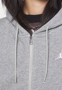 Sudadera gris con cremallera hecha de un tejido suave. Presenta un logotipo blanco, cordones y una capucha ajustada. Cierre frontal con cremallera.