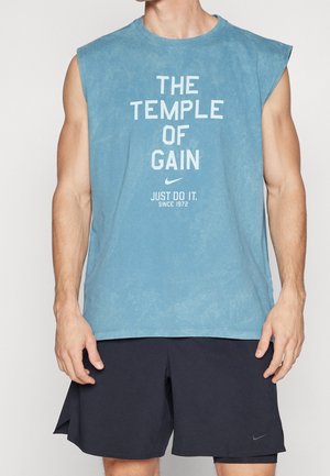 Muskularna osoba w niebieskiej koszulce bez rękawów z napisem "The Temple of Gain" i czarnych szortach, pozująca na jednolitym tle.
