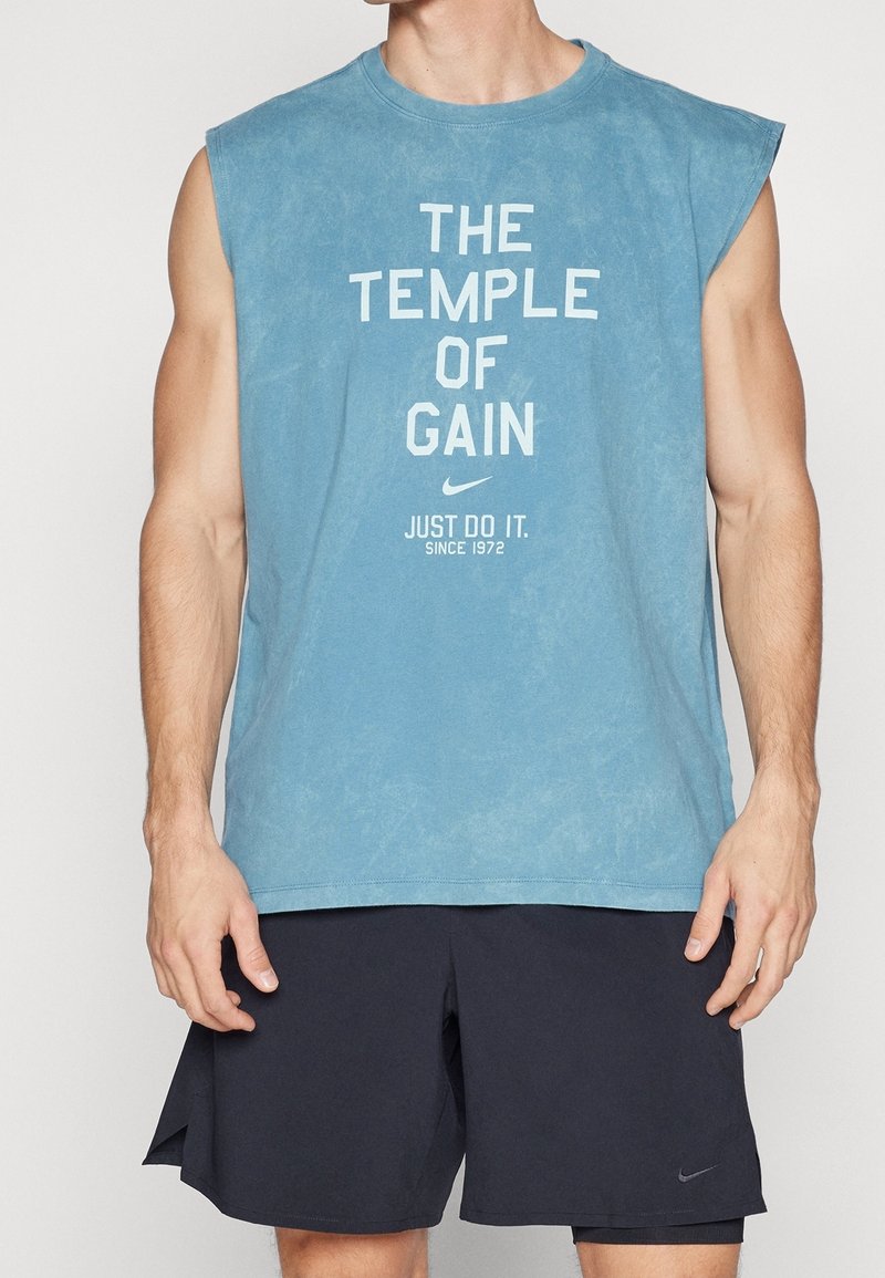 Personne musclée portant un débardeur bleu avec le texte « The Temple of Gain » et un short noir, posant devant un fond uni.