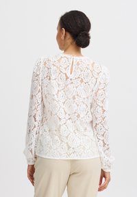 Hvid blondebluse med blomsterdesign, lange ærmer, rund hals med fint trim og en nøglehulldetalje bagpå.
