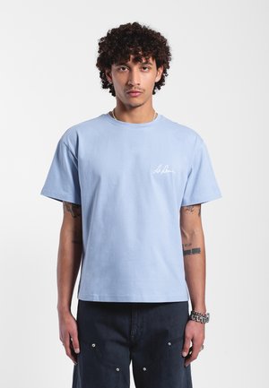 Camiseta de algodón de color azul claro con cuello redondo y mangas cortas. Presenta un logotipo bordado en blanco en el lado izquierdo del pecho, con un corte relajado y una textura suave.
